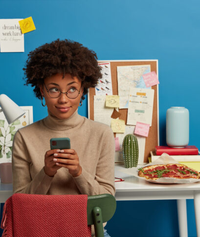Image of attractive curly haired female works remotely at home, holds mobile phone in hands, sends text messages, wears optical glasses, poses in own cozy room over workplace. People, work, technology