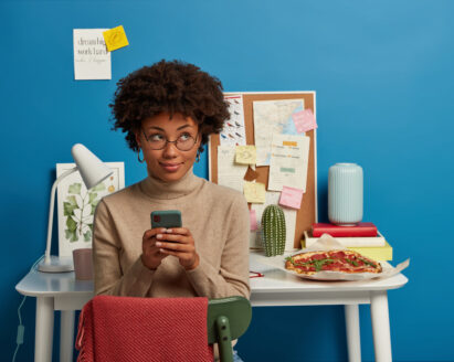 Image of attractive curly haired female works remotely at home, holds mobile phone in hands, sends text messages, wears optical glasses, poses in own cozy room over workplace. People, work, technology