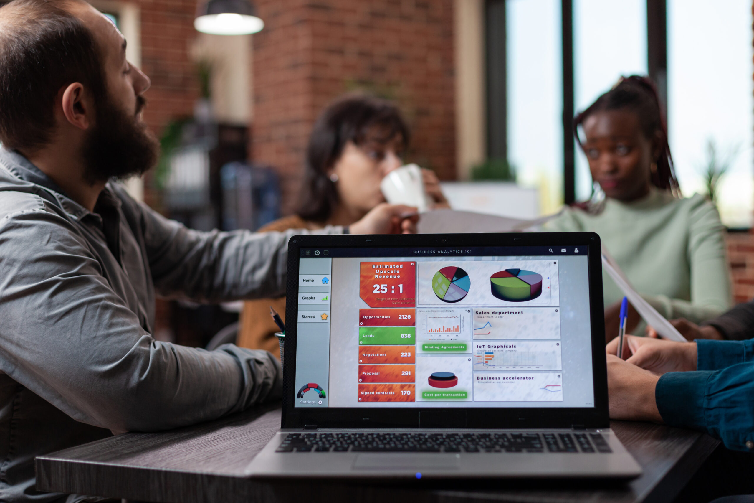 Laptop computer with company charts on screen standing on table in startup office while diverse team sharing papers with marketing turnover on it. Multiethnic businesspeople analyzing graphs