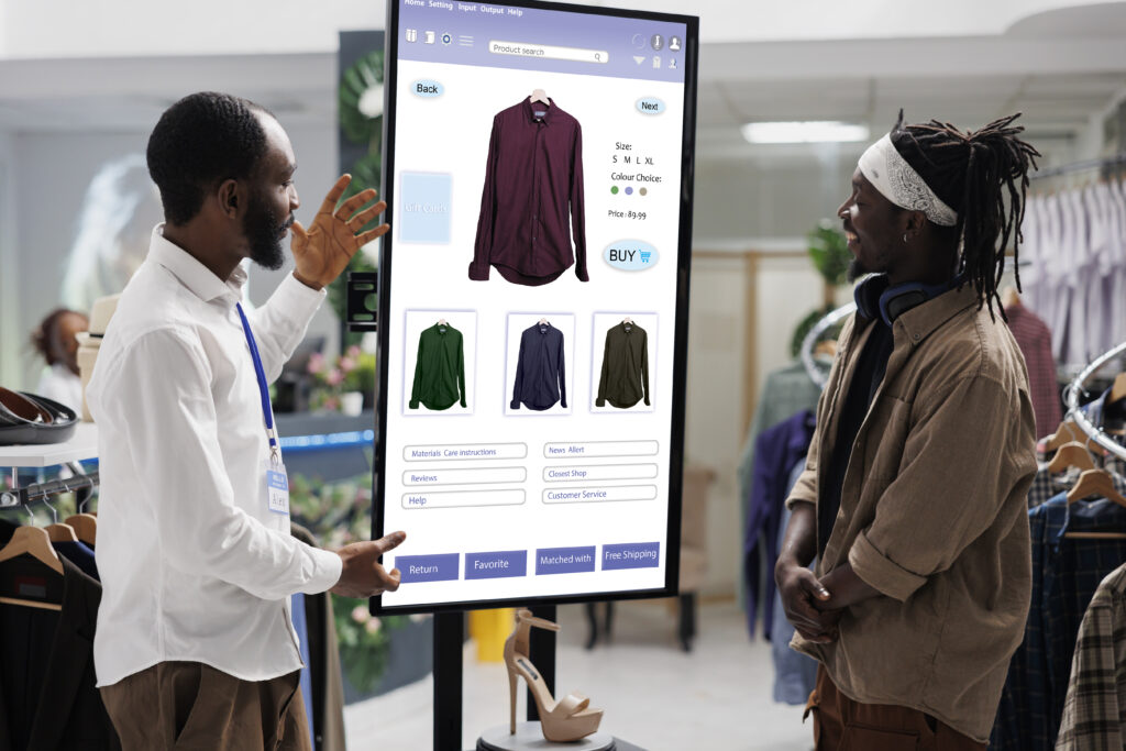 People look at clothing kiosk service with interactive board in fashion retail store. Male worker helping customer to buy modern clothes and items on self ordering touch screen monitor.